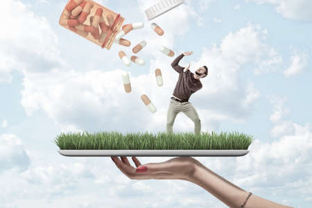 Womans hand holding digital tablet with green lawn on screen and little young man standing on it raising hands to protect from pills falling down from big jar.の写真素材