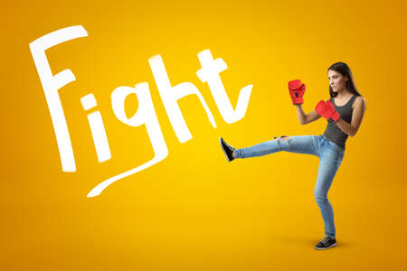 Side view of young pretty girl in jeans, sleeveless top and red boxing gloves with leg lifted for a kick on yellow background with Fight title.の写真素材