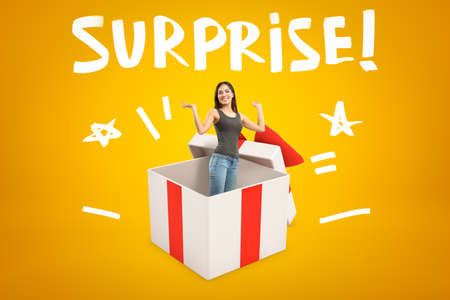 Young smiling woman in sleeveless top and blue jeans appearing from big white gift box on yellow background with title SURPRISEの写真素材