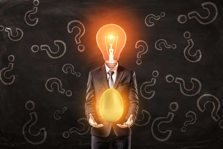 Man in suit with lightbulb instead head, levitating big shiny golden egg above palms, standing against black wall with pattern of question marks on it.の写真素材
