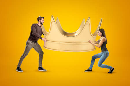 Side view of man and woman, both in casual clothes, carrying enormous gold crown on yellow background.の写真素材