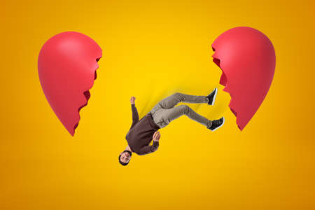 Young man in casual clothes falling between two broken heart pieces on yellow background