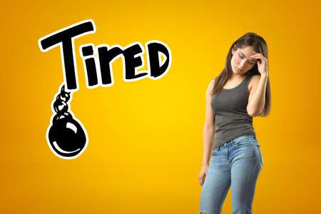 Young sad brunette girl wearing casual jeans and t-shirt with TIRED sign and black chained ball drawn on yellow backgroundの写真素材