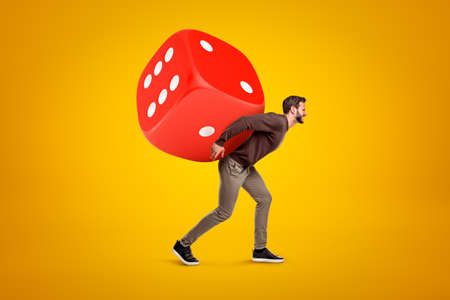 Young man in casual clothes carrying big red casino dice on his back on yellow backgroundの写真素材