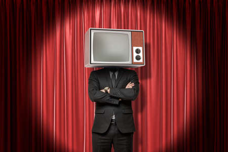 Front crop image of man in suit, with arms folded, and with TV set instead head, standing in spotlight at red stage curtain.の写真素材