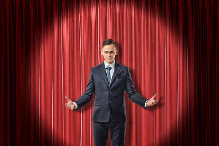 Young serious businessman with palms up on red stage curtains backgroundの写真素材