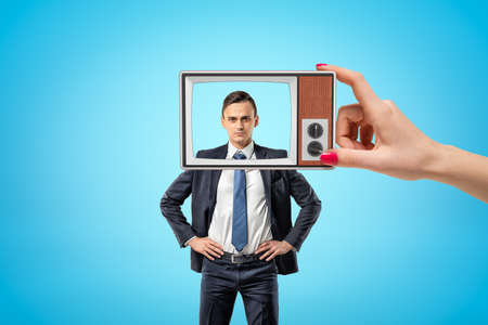 Young serious businessman standing, hands on hips, looking at camera through old TV frame with...