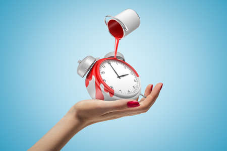 Female hand holding alarm clock with red paint bucket turned upside down above it on blue background