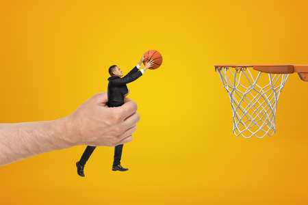 Big male hand holding businessman with basketball ball reaching out to hoop on yellow backgroundの写真素材