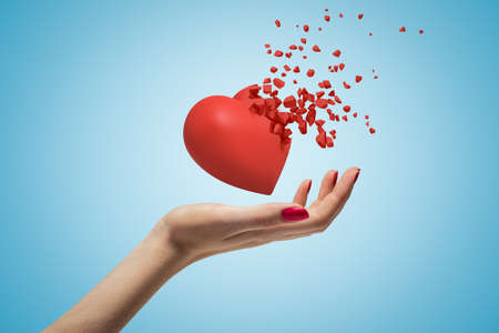 Close-up of womans hand facing up and levitating red valentine heart that has started to disintegrate into pieces on light-blue background.の写真素材
