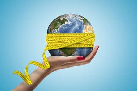 Female hand holding Earth globe covered with yellow measuring tape on blue background