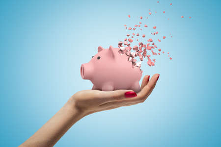 Close-up of woman's hand facing up and holding cute pink piggy bank that has started to disintegrate into pieces on light-blue background. Money worries. Financial difficulties. Profit loss.の写真素材