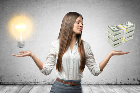 Crop image of young beautiful businesswoman, hands at sides, palms facing up and levitating light bulb and stack of cash.の写真素材