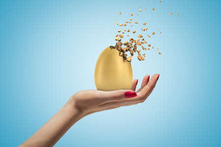 Female hand holding golden egg shattering into pieces on blue backgroundの写真素材
