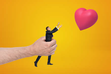 Big hand holding small businessman reaching out with his both hands for red valentine heart on amber background.の写真素材