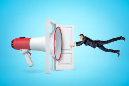 Side view of huge megaphone in open doorway knocking businessman off his feet with sound wave and making him grab at door so as not to fly away.の写真素材