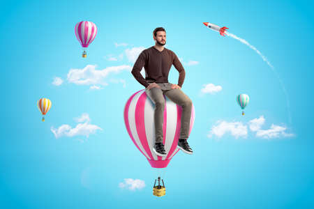 Serious young man sitting on top of pink and white hot air balloon in blue sky with other hot air...