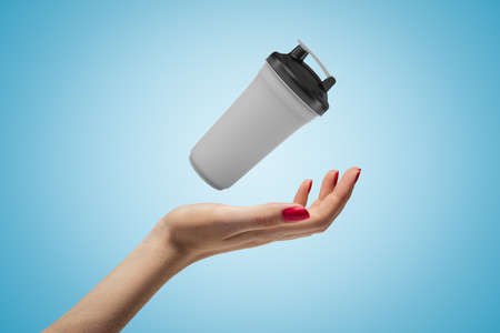 Close-up of womans hand facing up and levitating plastic sport cup on light-blue background.