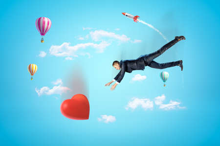 Businessman reaching to big red heart with hot air balloons and silver red space rocket in the air on blue backgroundの写真素材