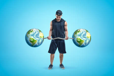 Young athlete in black cap, sleeveless hoodie and shorts, standing and looking down at barbell in...