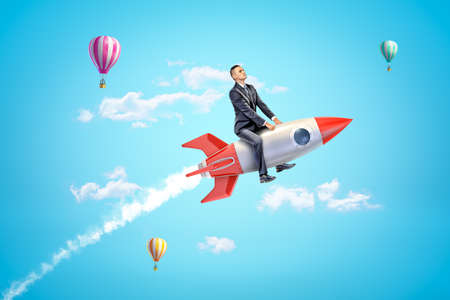 Young businessman riding toy rocket in blue sky with hot air balloons in background.の写真素材