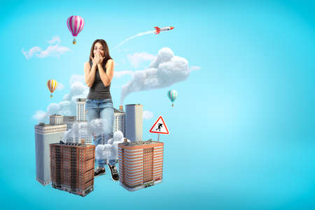 Scared giant young woman covering mouth with hands, standing amid high-rise buildings that are emitting smoke on blue background with copy space.の写真素材