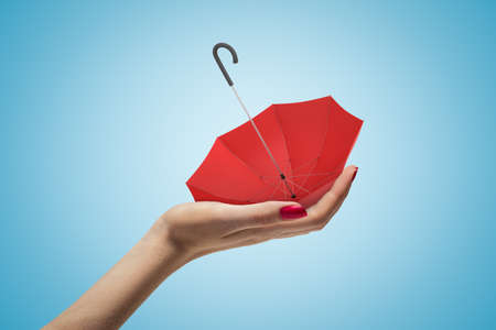 Side closeup of womans hand holding small red umbrella upside down on her palm on light blue gradient background.の写真素材