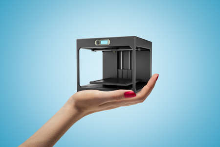 Side closeup of womans hand facing up and holding black 3D printer on light blue background.