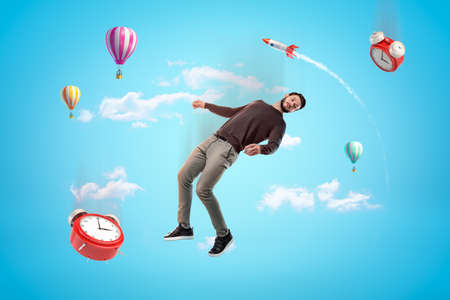 Young man and two alarm clocks floating in blue sky with rocket and hot air balloons in background.の写真素材