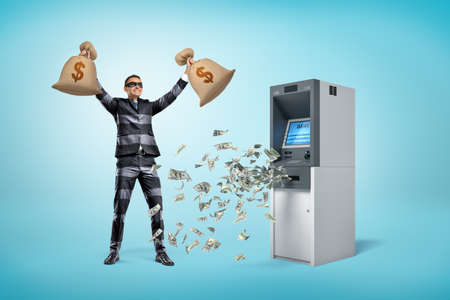Happy male robber holding money bags with dollars falling out of ATM machine on blue backgroundの写真素材