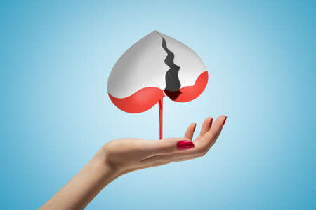 Side view of womans hand facing up and levitating gray broken heart upside down, with red liquid dripping down, on light blue background.の写真素材