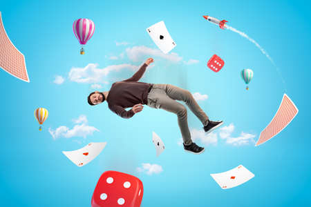 Man in casual clothes with red casino dice, playing cards, hot air balloons and silver red space rocket in the air on blue backgroundの写真素材