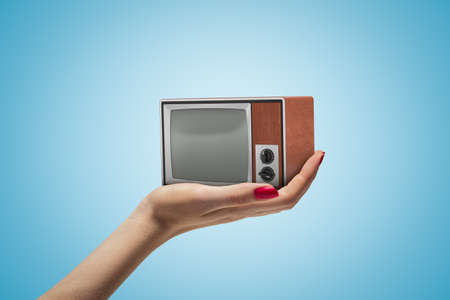 Side closeup of womans hand facing up and holding small retro TV set on light blue gradient background.の写真素材