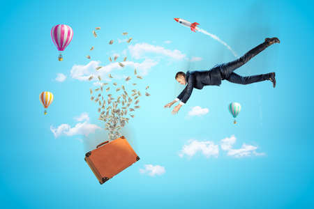 Businessman reaching to brown suitcase with dollars flying out, hot air balloons and silver red space rocket in the air on blue backgroundの写真素材