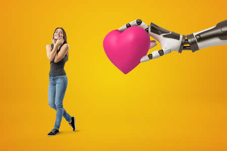 Young happy brunette girl wearing casual jeans and t-shirt and big robotic hand holding pink heart on yellow backgroundの写真素材