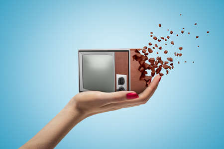 Female hand holding retro vintage TV set shattering into pieces on blue backgroundの写真素材