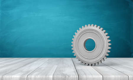 3d rendering of light-grey metal gearwheel standing on wooden floor near blue wall with some copy space on the left.の写真素材