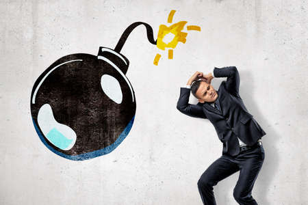 Young businessman protecting his head with his hands and big cartoon ball bomb on white background