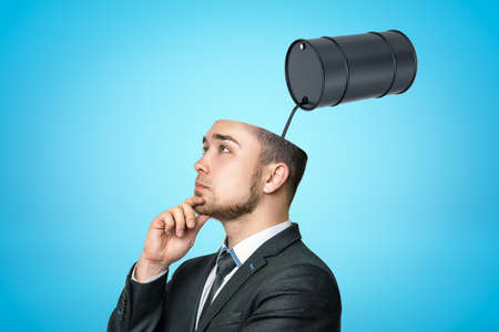 Thinking businessman with black thick liquid spilling out from black oil barrel in his head on blue backgroundの写真素材
