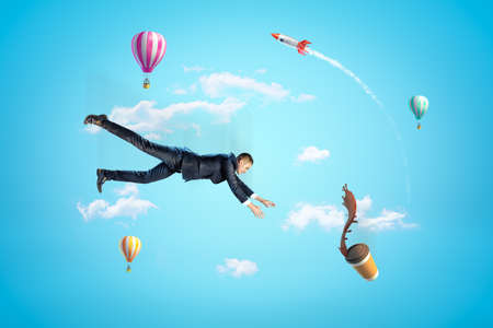 Young businessman in sky falling down trying to catch paper coffee cup which is spilling coffee in air with hot air balloons and rocket in background.の写真素材