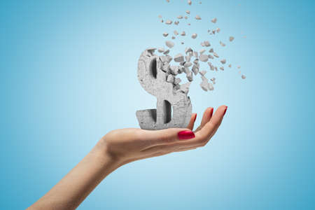 Female hand holding stone concrete dollar sign shattering into pieces on blue backgroundの写真素材