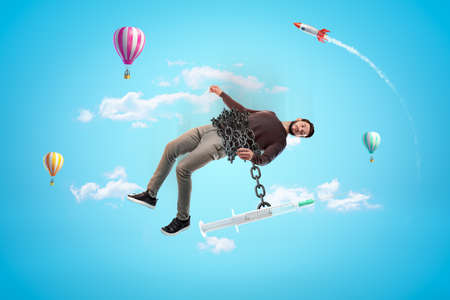 Man in blue sky with massive chain around waist with huge syringe hanging down on chain and pulling man down.の写真素材