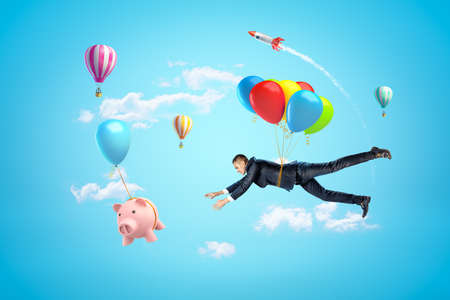 Businessman with colorful air balloons reaching to pink piggy bank with hot air balloons in the air on blue backgroundの写真素材