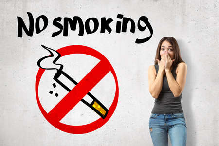 Crop image of young surprised girl covering mouth with hands, standing near wall with title No Smoking.の写真素材