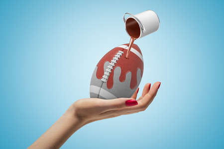 Female hand holding american football ball with small silver paint bucket turned upside down and brown paint pouring on it on blue backgroundの写真素材