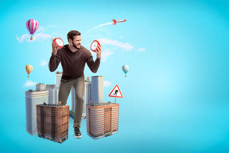 Angry young man in casual clothes holding two alarm clocks with city skyscrapers and hot air balloons on blue backgroundの写真素材