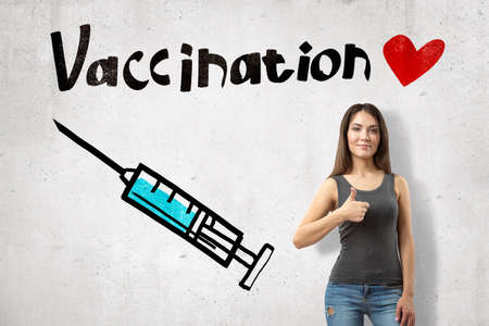 Young smiling girl giving thumbs up near wall with image of syringe and title Vaccination with cute little red heart.の写真素材