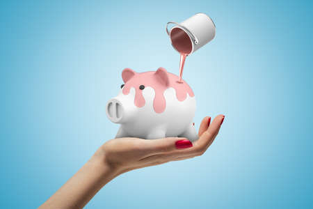 Closeup of woman's hand facing up holding white piggy bank and can of pink paint in air poring paint onto piggy on light-blue background. Build business. Monetize talent. Make money.の写真素材