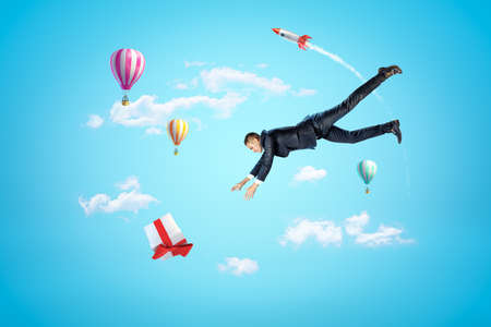Businessman reaching to gift box with hot air balloons and silver red space rocket in the air on blue backgroundの写真素材