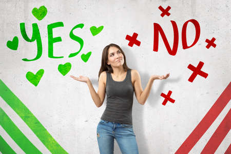 Young woman in casual clothes showing doubt with Yes and No sings on white backgroundの写真素材